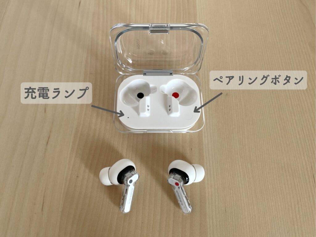 Nothing Ear(a)のケースからイヤホンを出してある。右イヤホンに赤丸、左イヤホンに黒丸が付いており、ケースのしまう場所にも対応した赤丸と黒丸のマークが付いている。ケースの右下にペアリングボタンがあり、左下に充電状況を示すランプがある。