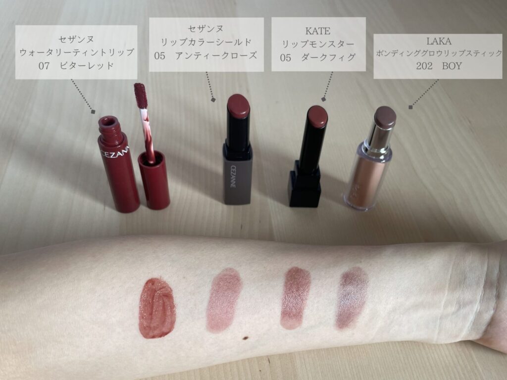口紅4本を並べ、それぞれを腕にスウォッチしている。左から、セザンヌのウォータリーティントリップの07ビターレッド、セザンヌのリップカラーシールドの05アンティークローズ、毛糸のリップモンスターの05ダークフィグ、ラカのボンディンググロウリップスティックの202ボーイ。