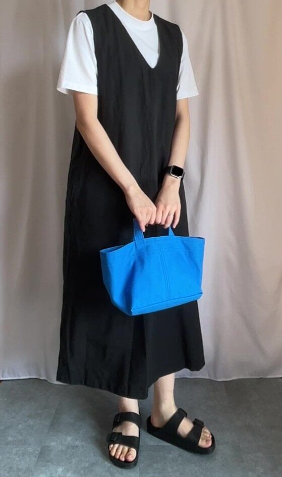 白いTシャツに黒いジャンパースカートを着て、鮮やかなブルーのトートバッグを持っている。