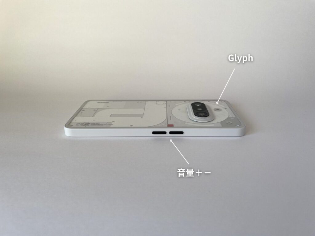 NothingのPhone(3a)を伏せて置いてある。サイドに音量ボタンがあり、カメラの周りにグリフのライトがあることを図示している。